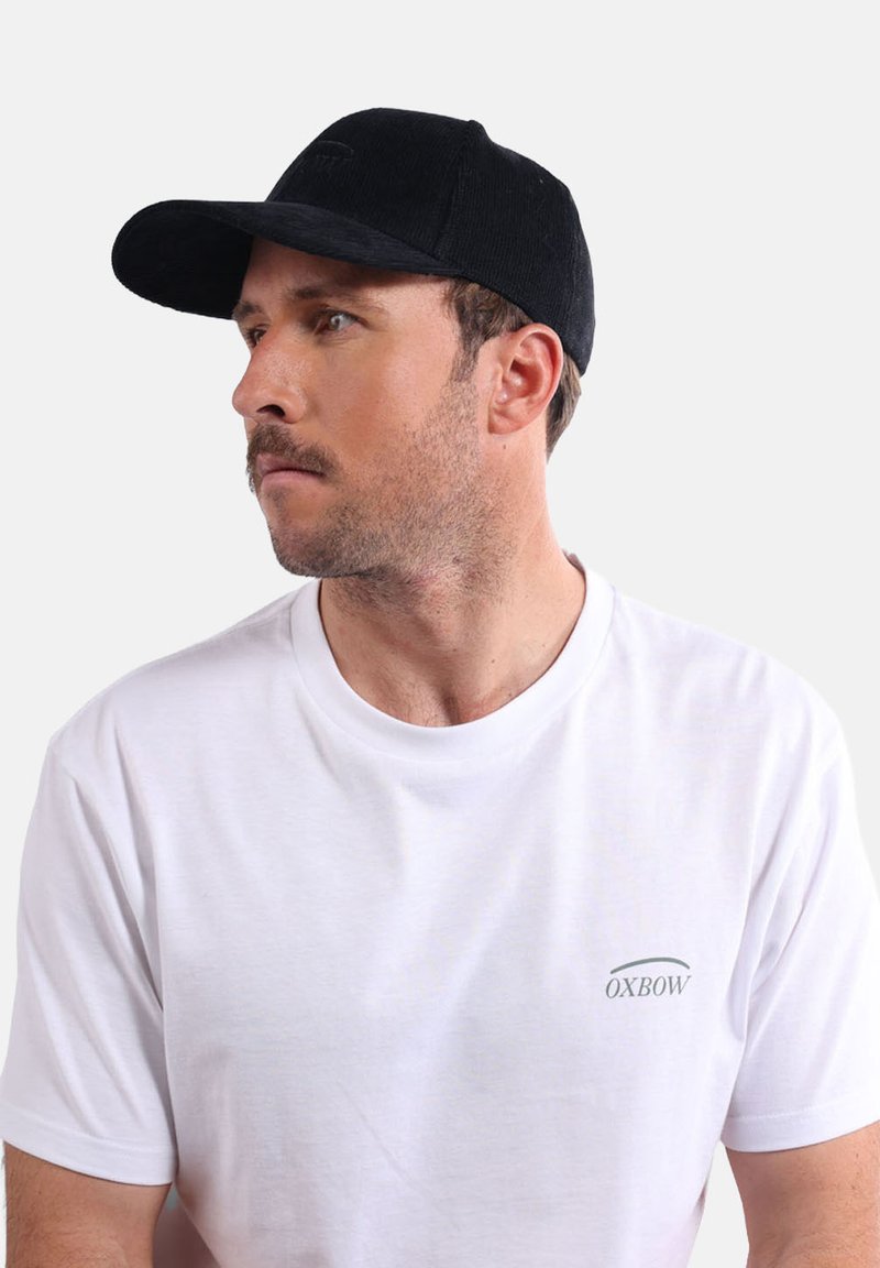 Casquette noire avec un bord incurvé et un tissu texturé, portée avec un t-shirt blanc uni comportant un petit logo gris sur la poitrine.