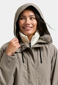 Vesitiivis beige takki, jossa on huppu, fleecevuorattu kaulus ja säädettävät nyörit. Varustettu napillisiin kiinnityksiin ja näkyviin ompeleisiin.