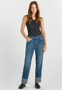 Jeans en denim bleu taille haute avec ourlets retroussés, comprenant cinq poches et une fermeture à boutons. Portés avec un débardeur noir et des chaussures noires.