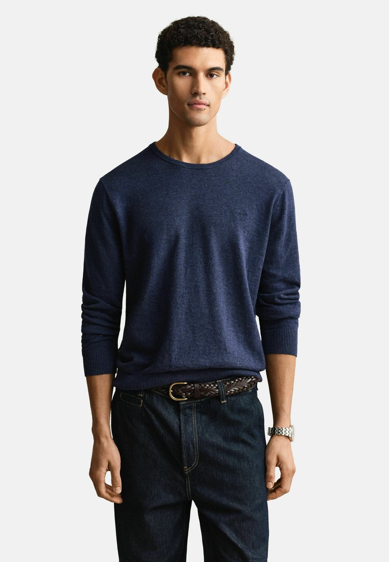 Homme portant un pull à manches longues bleu marine, un jean foncé avec une ceinture tressée, et une montre-bracelet argentée, debout devant un fond blanc.