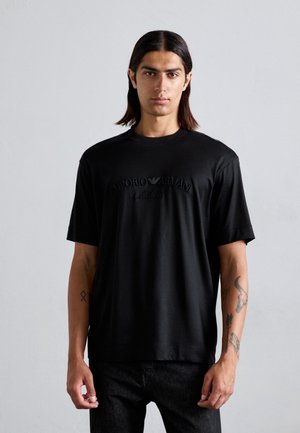 Svart t-shirt med kort ärm och rund halsringning, med ett präglat "EMPORIO ARMANI Milano"-logotyp på bröstet. Slätt tyg och avslappnad passform.