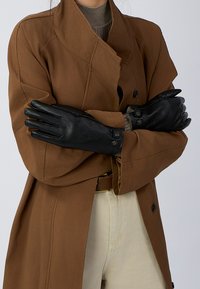 Zwarte leren handschoenen met een gladde textuur, voorzien van een knoopsluiting bij de manchet, gedragen over een bruine jas en beige broek.