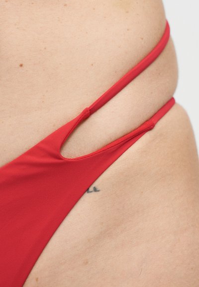 Κοντινό πλάνο στο ισχίο ενός ατόμου που φορά κόκκινο κάτω μέρος μαγιό με λεπτές τιράντες και ένα μικρό μερικό τατουάζ ορατό στο δέρμα.
