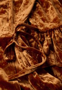 Tissu en velours d'un brun riche avec une texture douce. Présente une ceinture nouée à la taille et des détails froncés le long du bord.