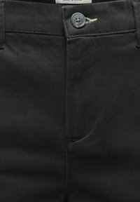 Pantalon noir avec ceinture boutonnée, braguette zippée, passants pour ceinture et poche avant, présenté en vue rapprochée.