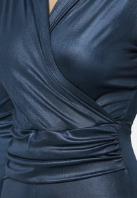 Marineblauwe wikkeljurk van glad, glanzend materiaal; beschikt over geplooide zijdetails en een diepe V-halsontwerp met subtiele textuur.