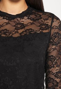 Top in pizzo nero con motivi floreali, colletto alto, maniche lunghe in tessuto trasparente e una sezione in tessuto nero solido nella parte inferiore.