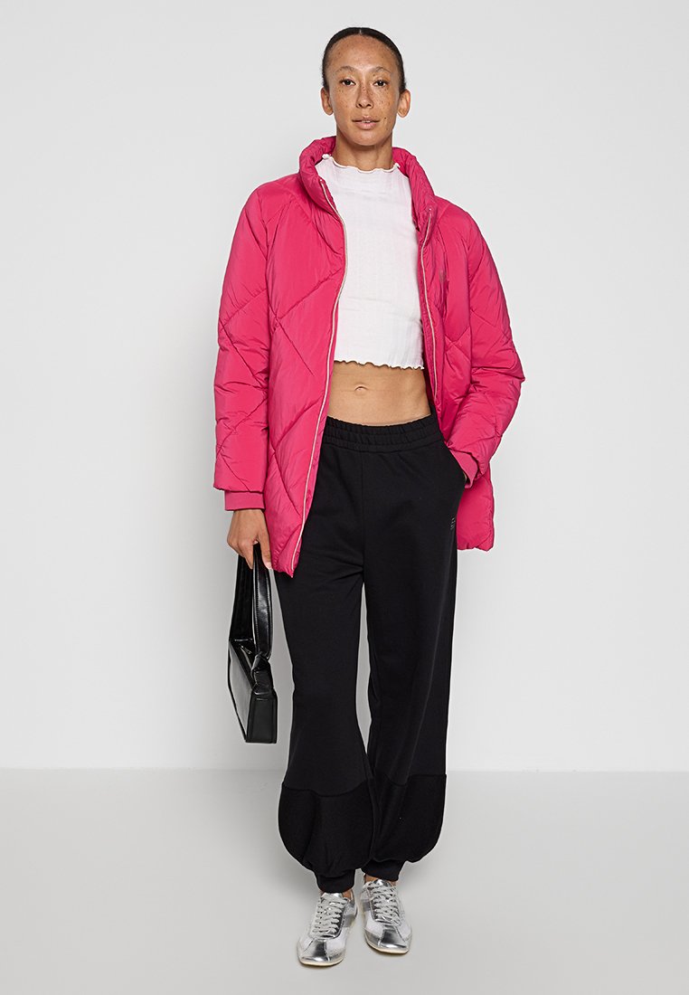 Fuchsia gesteppte Pufferjacke mit Kapuze, verkürztes weißes Oberteil, schwarze lässige Hose mit Bündchen und silberne Sneakers. Schwarze Handtasche.