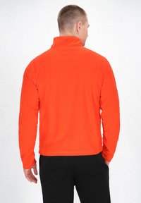 Orange fleecejakke med høj krave, lange ærmer og en glat tekstur, sammen med sorte bukser. Har en lige kant.
