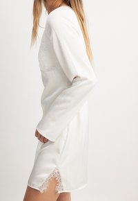 Žena v bílé saténové šatech s krajkovými detaily na výstřihu a lemu, stojící bokem proti jednobarevnému pozadí.