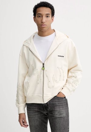 Felpa con zip crema, decorata con un ampio motivo ricamato in perline bianche sulle maniche e sul davanti, dotata di cappuccio e polsini a costine. Accento del logo nero.