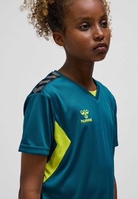 Jeune personne aux cheveux bouclés portant un maillot de sport bleu sarcelle avec des accents jaunes et un logo hummel, debout devant un fond uni.