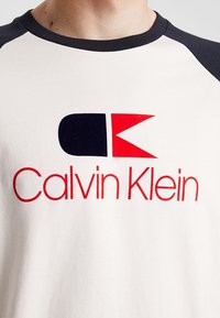 Camiseta de algodón blanca con mangas raglán azul marino. Presenta un bold logotipo en rojo y negro de "Calvin Klein" y un emblema estilizado en la parte frontal.