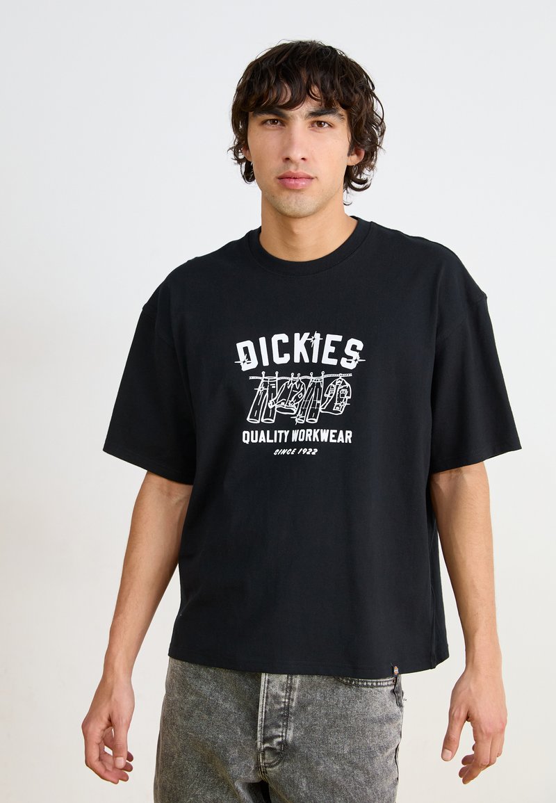 Juodas medvilninis marškinėlis su trumpomis rankovėmis, kuriame yra baltas grafikų atspaudas su užrašais "DICKIES" ir "QUALITY WORKWEAR". Matomos pilkos džinsinės kelnės.