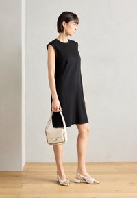 Vestito nero senza maniche con una vestibilità ampia, abbinato a una piccola borsa bianca e scarpe bianche con accenti metallici dorati. Sfondo in parquet.