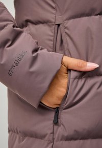 Giacca imbottita di un viola scuro con una mano nella tasca foderata in pile. Presenta una cerniera e il nome del marchio "67° N'ahk'ä" stampato sulla manica.