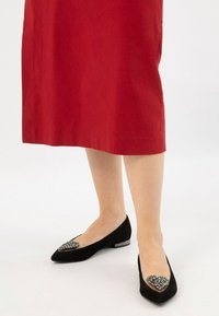 Gambe che indossano ballerine nere a punta con decorazioni di gioielli abbinate a una gonna rossa fino al ginocchio su uno sfondo bianco.