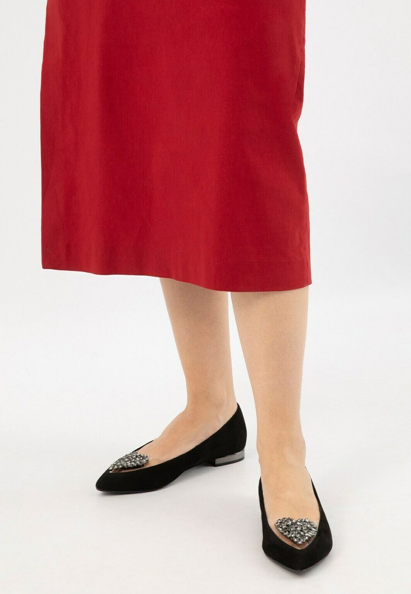 Gambe che indossano ballerine nere a punta con decorazioni di gioielli abbinate a una gonna rossa fino al ginocchio su uno sfondo bianco.