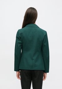 Blazer vert sur mesure avec col cranté, silhouette structurée et deux poches latérales. Texture lisse, coupe ajustée et lignes épurées.