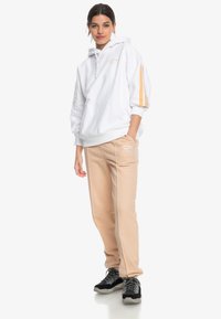 Felpa bianca con una tasca frontale e strisce arancioni sulle maniche, abbinata a pantaloni della tuta beige con un logo e una vita elasticizzata.