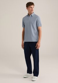 Cămașă polo cu dungi de culoare albastru deschis, cu mâneci scurte, purtată cu pantaloni navy și adidași albi. Are un guler și un singur nasture.
