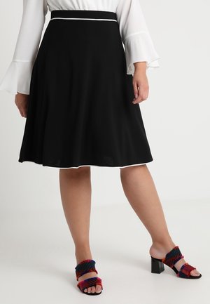 Kobieta w czarnej spódnicy do kolan z białą lamówką, białej bluzce z rozkloszowanymi rękawami oraz kolorowych, tkanych sandałach na obcasie.