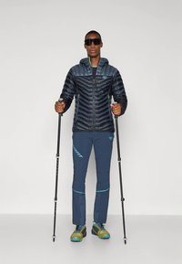 Szigetelt, sötétkék dzseki cipzárral és kapucnival; tengerészkék nadrág világoskék részletekkel. A felhasználó túrabotokkal és napszemüveggel.