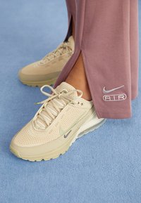 Beige Nike Air sneakers with mesh details worn with mauve pants featuring Nike Air logo, standing on light blue carpet.