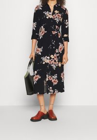 Květinové midi šaty v černé barvě s růžovými a oranžovými květy. Mají límečkový výstřih, tříčtvrteční rukávy a zavázaný pas. Doplněny hnědými dřeváky.