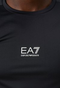 Svart treningsskjorte laget av glatt, elastisk materiale. Har en hvit EA7-logo med "EMPORIO ARMANI"-tekst under, kroppsnær design.