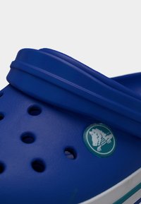 Κοντινό πλάνο μπλε λαστιχένιου παπουτσιού τύπου κλογκ με οπές αερισμού και λουράκι, που φέρει ένα μικρό κυκλικό λογότυπο με έναν κροκόδειλο.