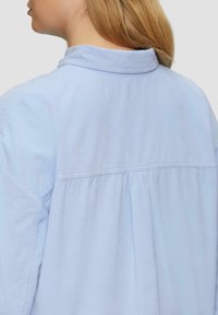 Lichtblauw, langmouw shirt gemaakt van zachte stof; heeft een puntige kraag en plooien aan de achterkant, wat een ontspannen pasvorm en een soepele textuur laat zien.