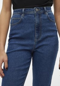 Modre jeans z visokim pasom, z zasnovo petih žepov, z zadrgo in kontrastnim šivom. Gladka tekstura in trpežno blago.