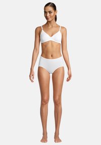 Frau steht barfuß und trägt einen weißen kabellosen BH und hochgeschnittene Unterwäsche vor einem schlichten weißen Hintergrund.