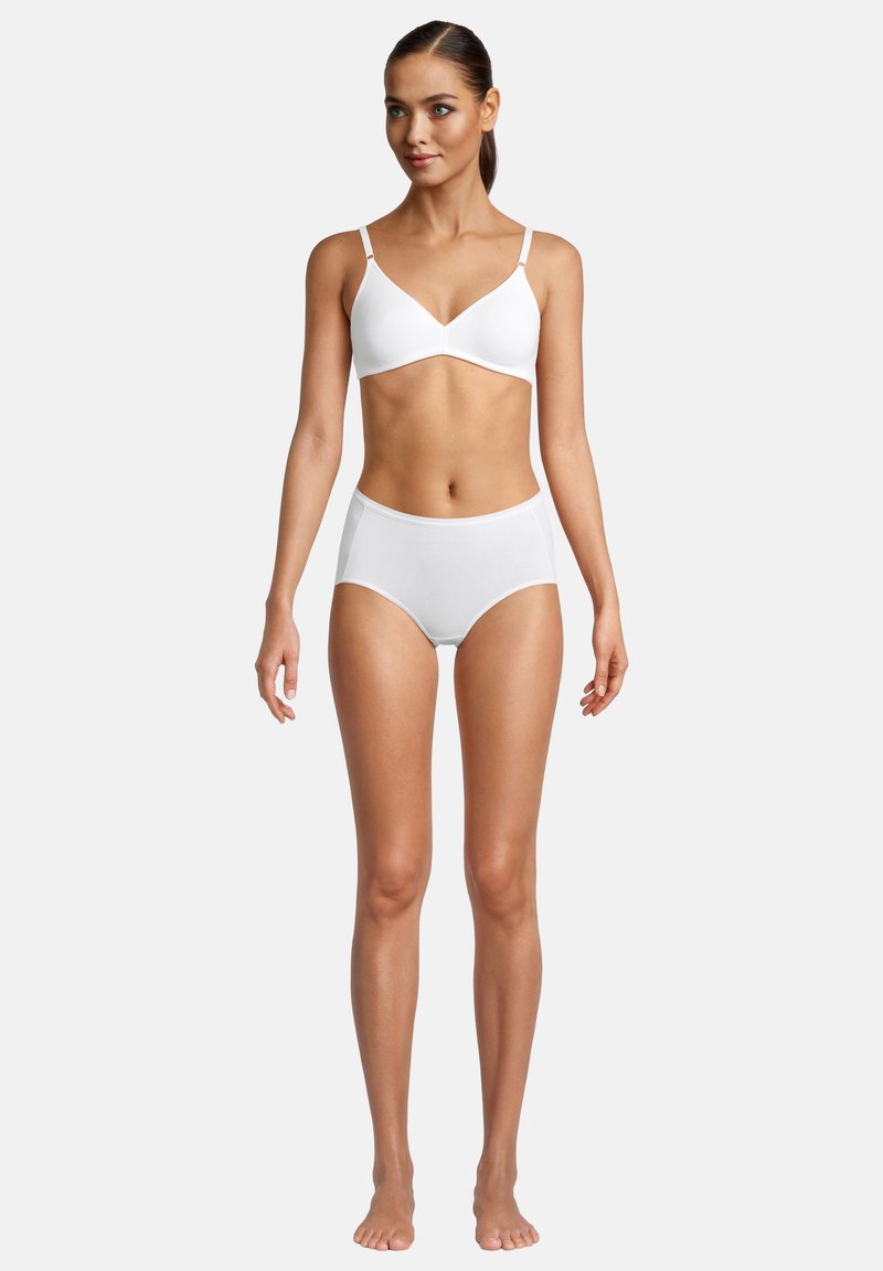 Frau steht barfuß und trägt einen weißen kabellosen BH und hochgeschnittene Unterwäsche vor einem schlichten weißen Hintergrund.