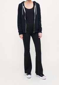 Sudadera negra con cremallera y cuerdas blancas, combinada con una camiseta sin mangas negra y pantalones de campana negros de cintura alta. Zapatillas negras con suelas blancas.