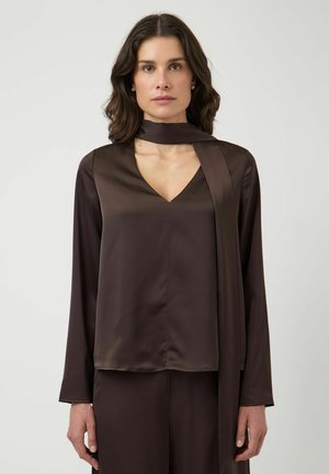 Mujer con cabello oscuro de longitud media lleva una blusa de satén marrón oscuro con cuello en V y mangas largas, acompañada de una bufanda larga a juego, sobre un fondo liso.