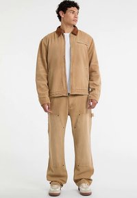 Zandkleurig katoenen jack met een bruine kraag en een voorzak, gecombineerd met zandkleurige cargobroek met zijzakken en een losse pasvorm.