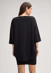Tunique noire surdimensionnée avec un ourlet arrondi et des manches amples, dotée d'un détail de bouton dans le dos. Fabriquée en tissu doux et lisse.