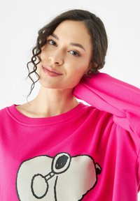 Rosa Sweatshirt mit einem großen Grafik eines Cartoon-Hundes mit Brille auf der Vorderseite. Der Stoff wirkt weich mit einer strukturierten Oberfläche.