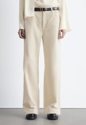 Beige Cordhosen mit weit geschnittenem Bein und glatter Textur, ausgestattet mit einem schwarzen Ledergürtel und Seitentaschen. Flacher Frontschnitt und entspannter Sitz.