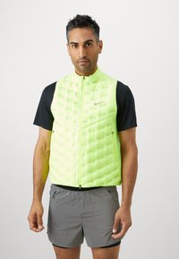 Gilet puffer Nike di un vivace verde lime con chiusura a zip, design trapuntato e tasche laterali, indossato sopra una maglietta nera a maniche corte.