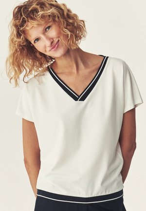 Blonde vrouw met krullend haar die een witte korte mouwen top draagt met een zwart gestreepte V-hals en zoom, glimlachend met het hoofd schuin.