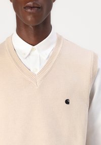 Μπεζ πλεκτό γιλέκο με σχέδιο V-neck, πάνω από λευκό πουκάμισο με κουμπιά. Διαθέτει μαύρο λογότυπο στην αριστερή πλευρά του στήθους. Λείος υφασμάτινος ιστός.