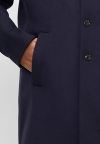 Cappotto di lana blu navy con design monopetto, due bottoni e tasca laterale, con una texture liscia e un taglio classico.