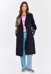 Čierny trenčkot s rukávmi v leopardom vzore, dvojradový dizajn, prehodený cez sveter s ružovými pruhmi a svetlomodré džínsy so zelenými akcentmi.