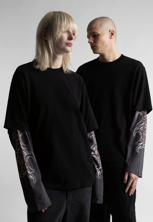 Dwie osoby ubrane w czarne koszulki z krótkim rękawem na długie rękawy z tatuażowym nadrukiem, stojące na tle jasnego, jednolitego tła i patrzące w różne strony.