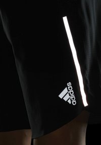 Pantaloni corti neri realizzati in materiale leggero, con una striscia riflettente sul lato e un logo Adidas argenteo vicino all'orlo. Design testurizzato.
