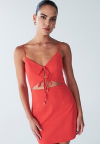 Punane lõhikuga kleit spagetiribadega, millel on V-kaelus ja ees lipsudetail. Valmistatud siledast kangast, millel on metallikaktsendid.