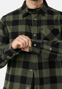 Groen en zwart geruite flanellen blouse met knoopsluiting, lange mouwen en een enkele borstzak. Voorzien van metalen drukknoopjes.